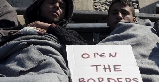 Dos refugiados descansan en el suelo cerca del pueblo de Idomeni, Grecia, con un cartel que pone 'abre las fronteras'./REUTERS
