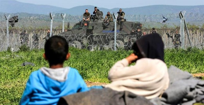 Varios niños descansan junto a la valla fronteriza mientras la policía macedonia hace guardia al otro lado cerca del campamento de refugiados de la ciudad de Idomeni. - EFE