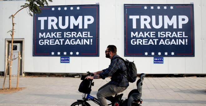 Un hombre montado en bicicleta pasa junto a unos letreros en Tel Aviv parafraseando el lema de campala de Donadl Trump. REUTERS/Baz Ratner