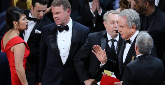 Los socios PricewaterhouseCoopers (PwC), Martha Ruiz y Brian Cullinan (a la izquierda de la imagen), conversan en el escenario de la gala de los Premios Oscar de Hollywood, mientras un regidor conversa con el actor Warren Beatty, quien sostiene uno de los