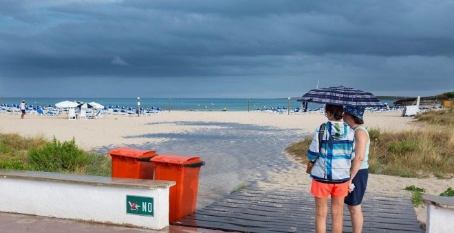 Dos mujeres se resguardan de la lluvia bajo un paraguas en una playa de Menorca | EFE/Archivo