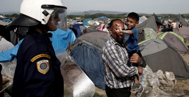 Un agente antidisturbios junto a un padre y su hijo en uno de los campamentos de refugiados improvisados en Idomeni, Grecia. - EFE