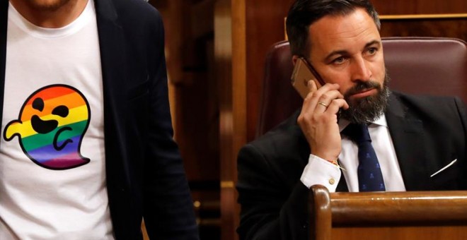 El presidente de Vox, Santiago Abascal, en uno de los escaños del Congreso de los Diputados, mientras pasa uno de los diputados del PSOE con 'gaysper' en su camiseta. - EFE
