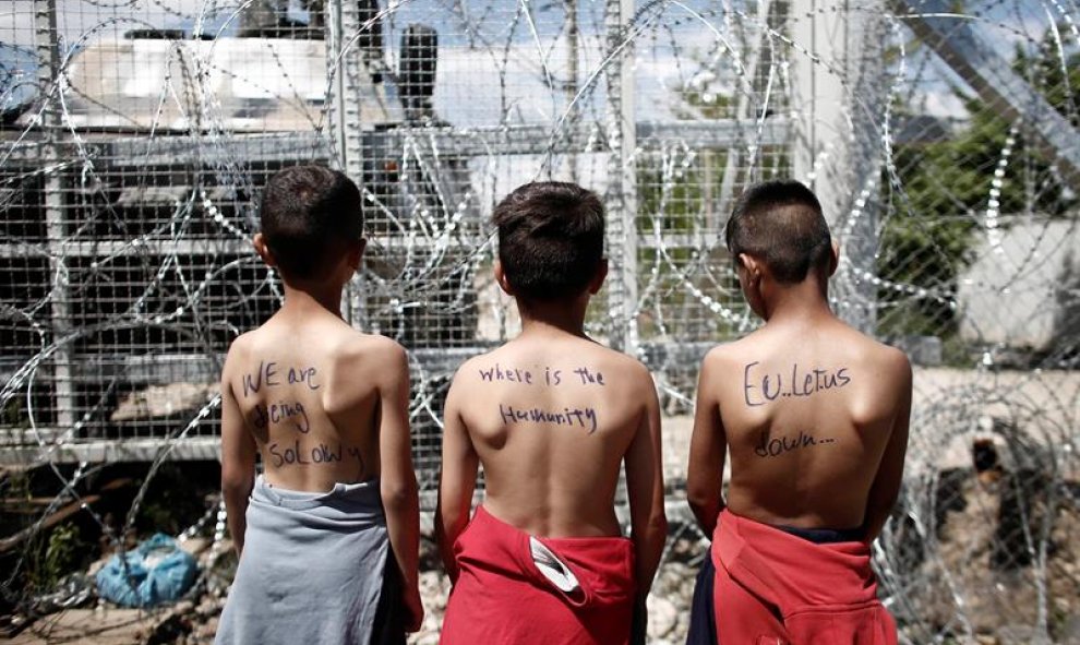 Un grupo de niños con mensajes en sus espaldas en los que se leen "Estamos muriendo lentamente", "¿Dónde está la humanidad?" y "La UE nos falla" participa en una protesta en el campamento de refugiados de Idomeni, Grecia. EFE/Kostas Tsironis