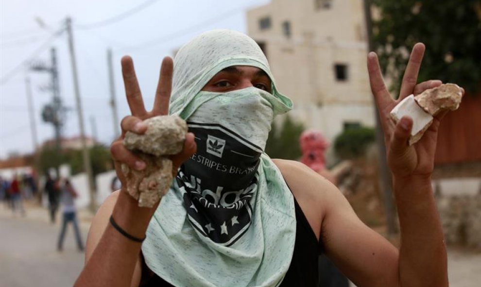 Un palestino con el rostro tapo, hace el gesto de la victoria con sus manos en las que lleva piedras, en la ciudad de Dura, cerca de Hebrón. /EFE-ABED AL HASHLAMOUN