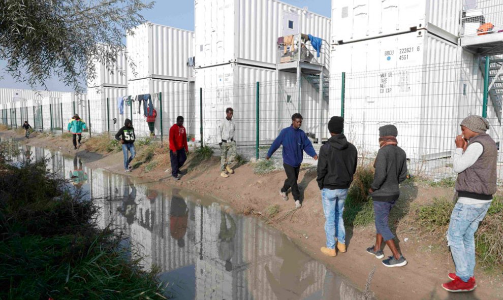 Varios inmigrantes se reflejan en el agua fangosa al pasar junto a los contenedores utilizados como vivienda improvisada, en la víspera del   desmantelamiento de La Jungla de Calais.- PHILIPPE WOJAZER (REUTERS)