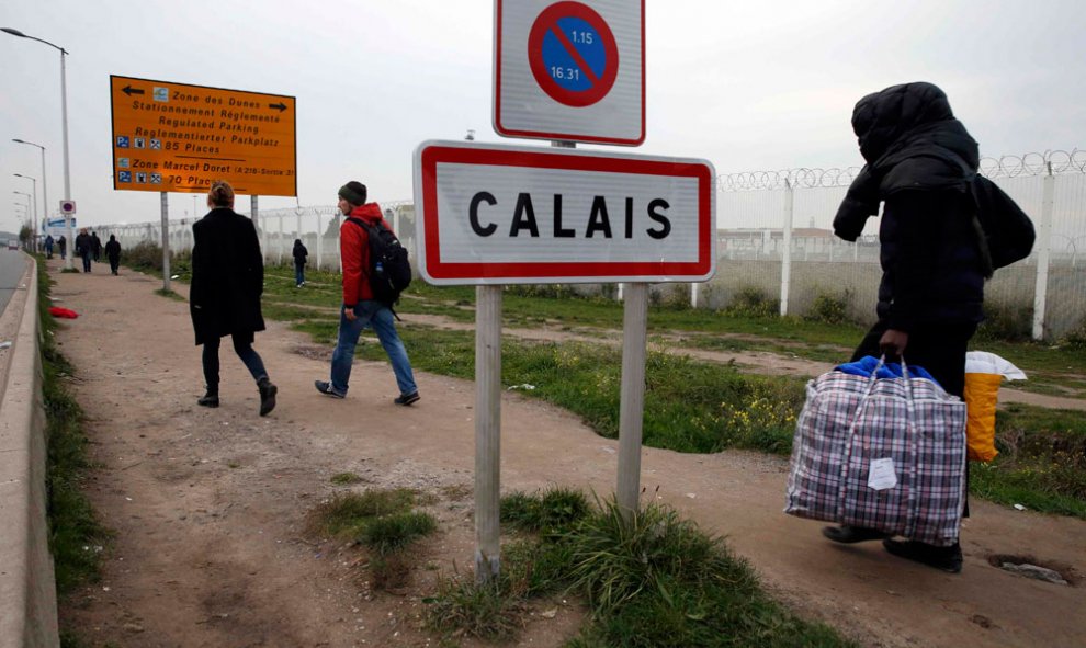 Un inmigrante traslada bosas con sus pertenencias junto a la señal que indica el límite urbano de la ciudad francesa de Calais.- PASCAL   ROSSIGNOL (REUTERS)