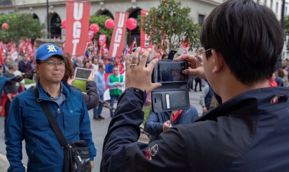 Unos turistas asiáticos se fotografían al paso de la manifestación del Primero de Mayo en Sevilla , donde miles de personas se han dado cita para reivindicar la igualdad de género, el empleo de calidad y las pensiones dignas, y cuyo lema es "Es tiempo de