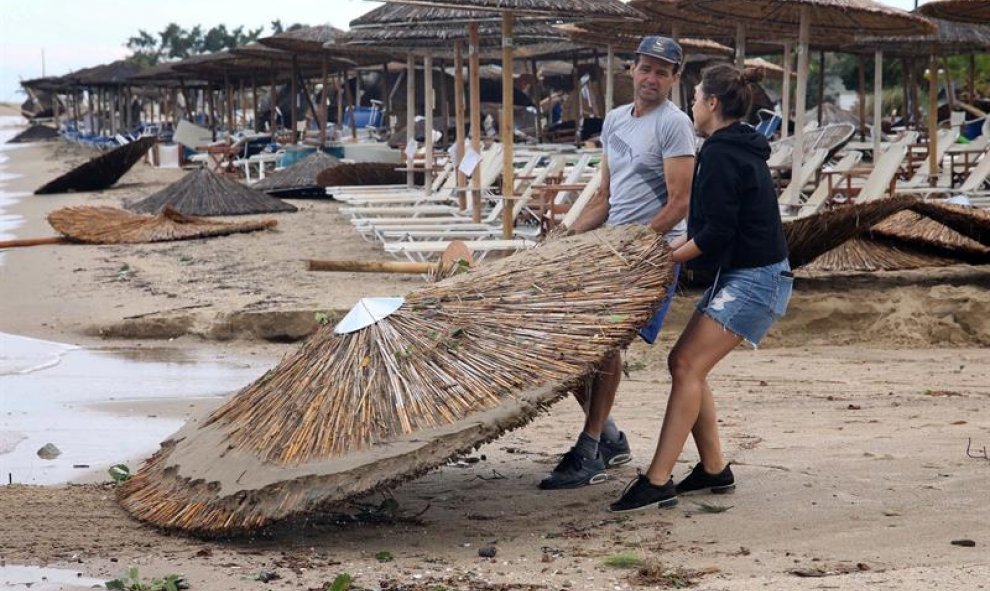 Dos empleados recogen las sombrillas que se han volcado por el temporal. EFE / VERVERIDIS VASSILIS