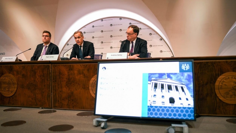 El actual gobernador del Banco de Inglaterra (c), Mark Carney, junto a su sucesor, Andrew Bailey (D), y el director de comunicación de la entidad, Mike Peacock (I), en una rueda de prensa en la sede de la institución para explicr las medidas adoptadas por