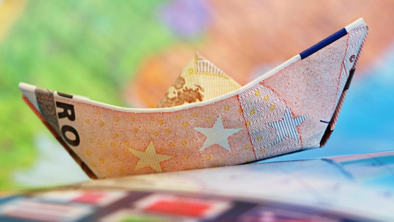 Imagen recurso de un barco de papel hecho con un billete. Al fondo, un mapamundi difuminado. - Pixabay