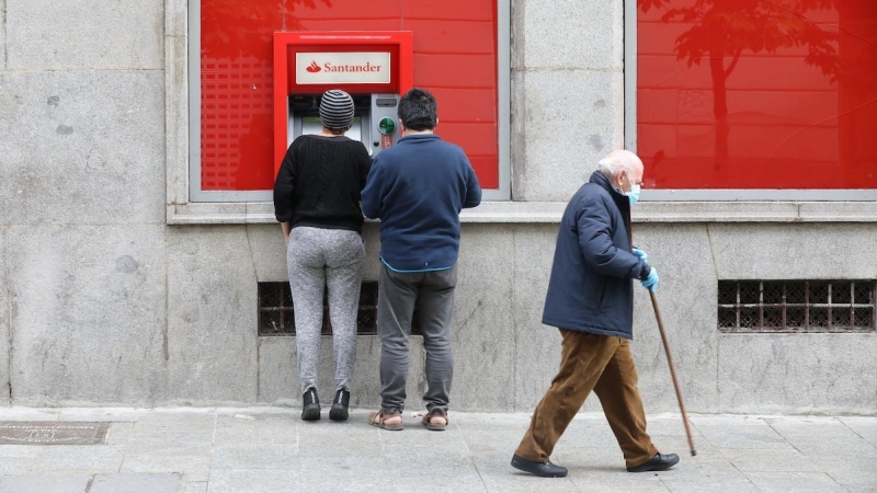 Un hombre con bastón camina por la capital al lado de dos personas que sacan dinero de un cajero, en Madrid, a 28 de abril de 2020.