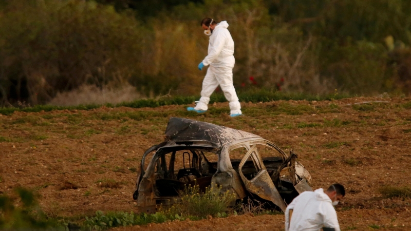 Expertos forenses caminan en un campo después de que una bomba explotara un automóvil (primer plano) y matara a la periodista de investigación Daphne Caruana Galizia en Bidnija, Malta, el 16 de octubre de 2017