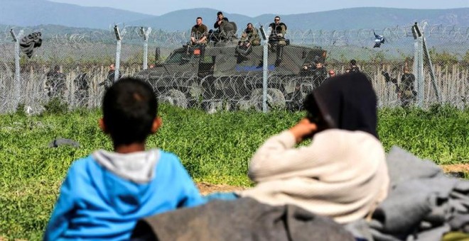 Varios niños descansan en Idomeni junto a la frontera, mientras la Policía macedonia hace guardia al otro lado de la valla. - EFE