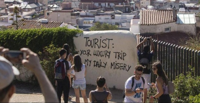 Turistas haciendo fotos cerca de una pintada en la que reza en inglés: 'Turista: tu viaje de lujo, mi miseria diaria'. | Quique García / EFE