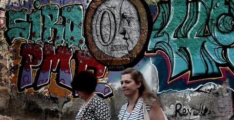 Dos mujeres caminan frente a un muro con un grafitti en el que aparece una moneda de euro con un 0 en su interior. AFP