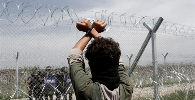 Un refugiado permanece tras la valla fronteriza frente a un grupo de policías macedonios durante una protesta en el campo de refugiados de Idomeni, situado en la frontera entre Macedonia y Grecia. EFE/Kostas Tsironis
