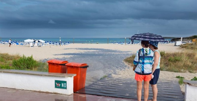 Dos mujeres se resguardan de la lluvia bajo un paraguas en la playa de Punta Prima, en el municipio de Sant Lluís (Menorca), en una jornada en la que la Agencia Estatal de Meteorología (Aemet) ha activado la alerta amarilla en Baleares por posibles precip