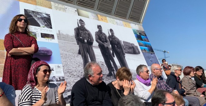 Assistents a la inauguració del mural en memòria dels afusellats al Camp de la Bota, davant de fotografia de soldats de l'exèrcit en pràctiques de tir en aquell mateix lloc. PÚBLIC