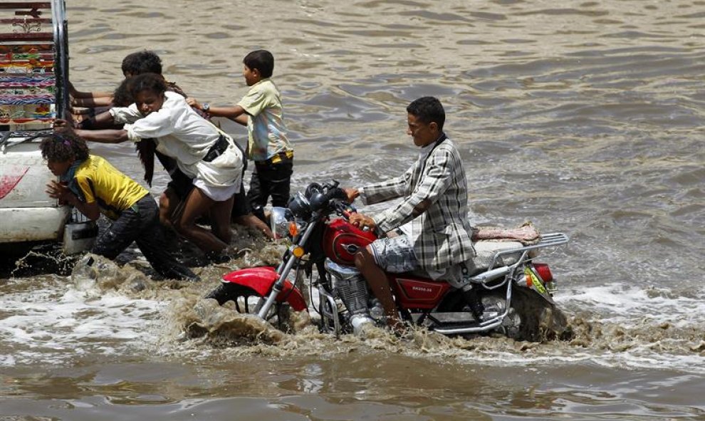 Un joven circula en motocicleta mientras varios niños empujan una camioneta en una calle inundada de Saná, Yemen. Intensas lluvias azotaron hoy varias regiones de Yemen causando importantes inundaciones. EFE/Yahya Arhab