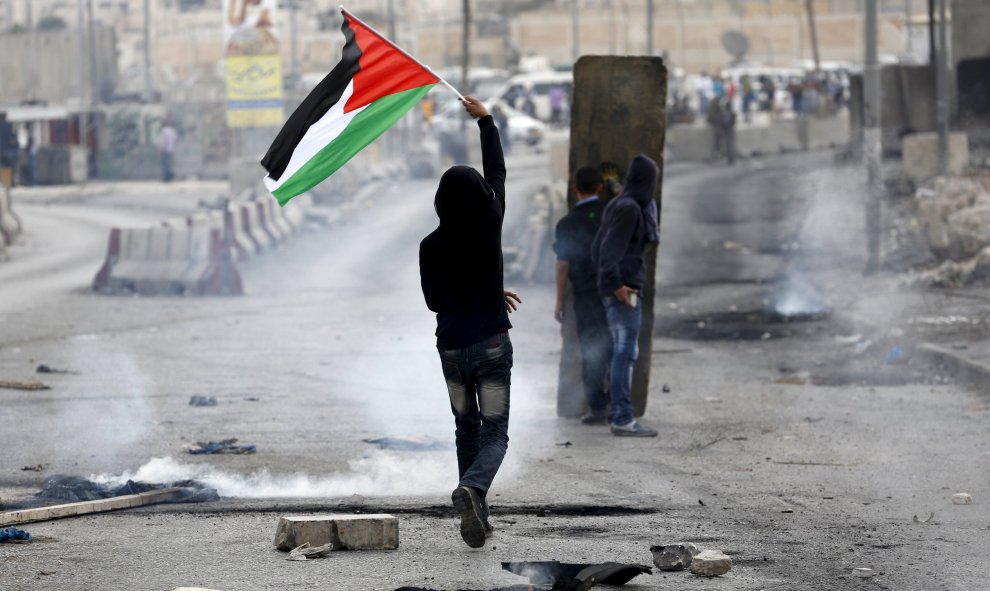 Un manifestante palestino sostiene una bandera palestina mientras otros se enfrentan con el ejército israelí en el puesto de control de Qalandia cerca de la ciudad ocupada de cisjordana, Ramala 6 de octubre de 2015. REUTERS / Mohamad Torokman