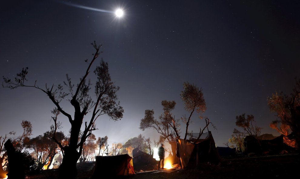 Un refugiado se calienta junto al fuego en un improvisado campamento cerca del centro de registro en la isla griega de Lesbos.- REUTERS / Yannis Behrakis