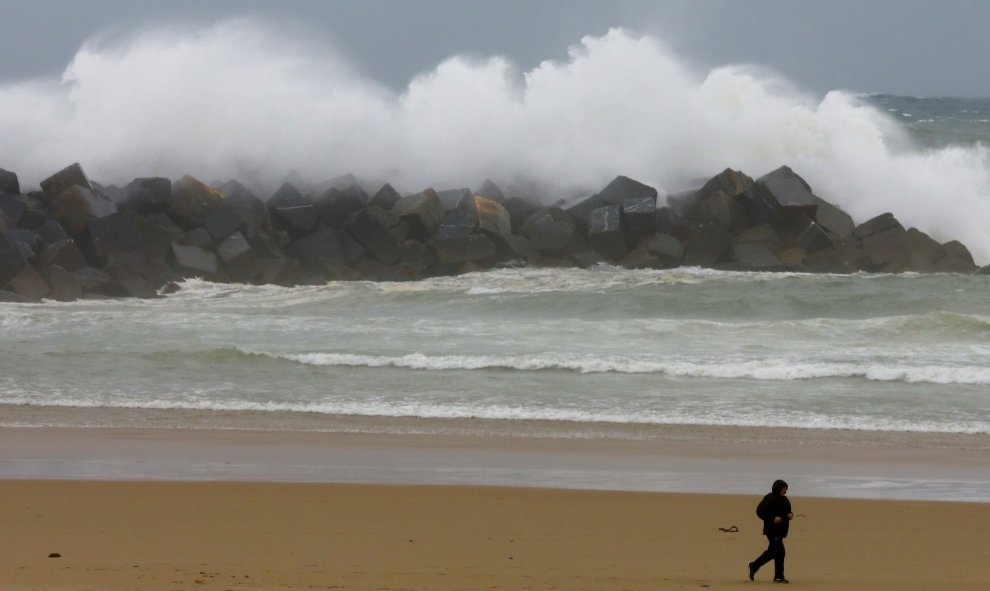 Un hombre se protege contra el viento y la lluvia en la playa de la Zurriola de San Sebastián, donde los cielos permanecen cubiertos con lluvias y chubascos generalizados. EFE/Juan Herrero.