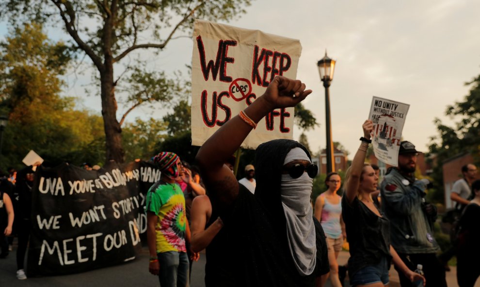 Un manifestante con el rostro tapado, encapuchado y con gafas de sol levanta el puño delante de una pancarta contra la represión policial | REUTERS