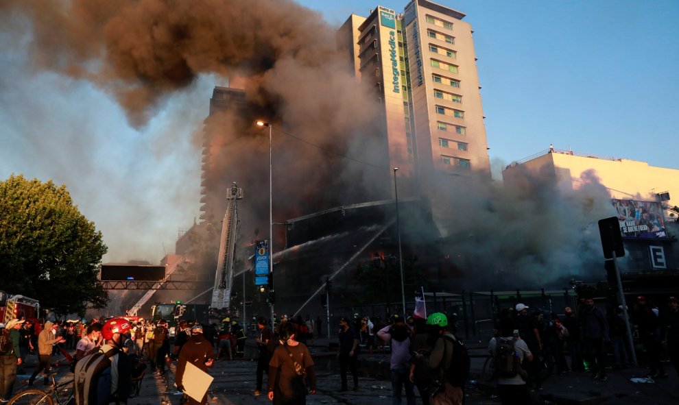 Los manifestantes frente a un edificio en llamas mientras los bomberos trabajan para apagar el fuego durante una protesta contra el gobierno en Santiag. REUTERS / Henry Romero