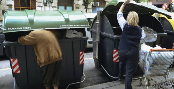 Una persona busca comida en un contenedor de basura de Bilbao, mientras una trabajadora de un supermercado echa bolsas a la basura. REUTERS