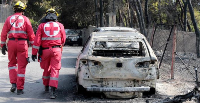 Miembros de la Cruz Roja caminan junto a un vehículo calcinado en una zona afectada por los incendios en Argyra Akti, en Mati. (PANTELIS SAITAS | EFE)