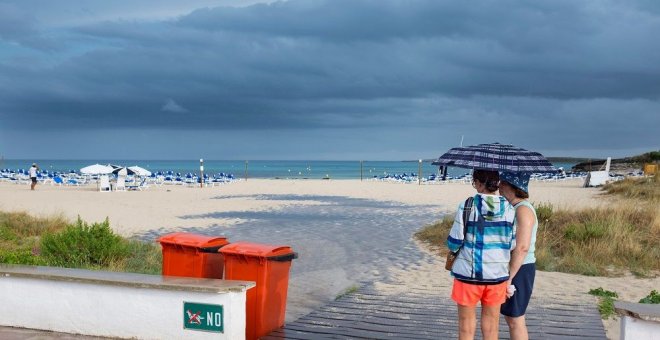 Dos mujeres se resguardan de la lluvia bajo un paraguas en una playa de Menorca | EFE/Archivo