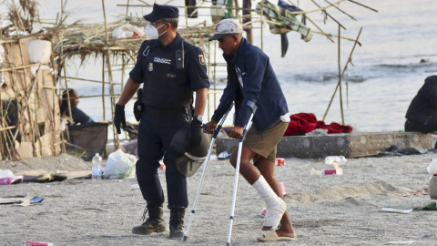 Un policía acompaña a uno de los migrantes, en muletas, durante el desalojo a pie de la playa del Trampolín en Ceuta, a 20 de agosto de 2026. Un policía acompaña a uno de los migrantes, en muletas, durante el desalojo a pie de la playa del Trampolín en Ceuta, a 20 de agosto de 2026.