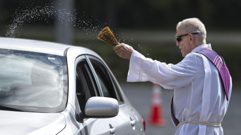 El padre Dennis Rausch rocía agua bendita sobre un coche durante un servicio religioso al que los fieles asisten en sus vehículos