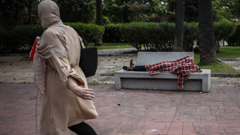 Una mujer observa a Mohamed, un menor marroquí de 17 años que duerme en el banco de los Jardines de Argentina de Ceuta tras entrar a nado a la ciudad a principios de esta semana.- JAIRO VARGAS Una mujer observa a Mohamed, un menor marroquí de 17 años que duerme en el banco de los Jardines de Argentina de Ceuta tras entrar a nado a la ciudad a principios de esta semana.- JAIRO VARGAS