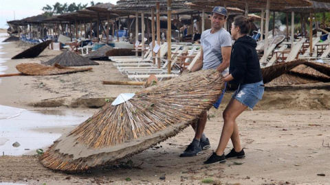 Dos empleados recogen las sombrillas que se han volcado por el temporal. EFE / VERVERIDIS VASSILIS