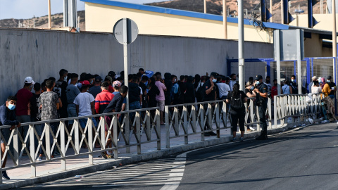 Agentes de Policía supervisan a cientos de personas, la mayoría marroquíes, que guardan cola en la oficina para tramitar los asilos situada en la frontera del Tarajal que separa Ceuta de Marruecos, a 3 de agosto de 2021, en Ceuta (España). Imagen de a Agentes de Policía supervisan a cientos de personas, la mayoría marroquíes, que guardan cola en la oficina para tramitar los asilos situada en la frontera del Tarajal que separa Ceuta de Marruecos, a 3 de agosto de 2021, en Ceuta (España). Imagen de a