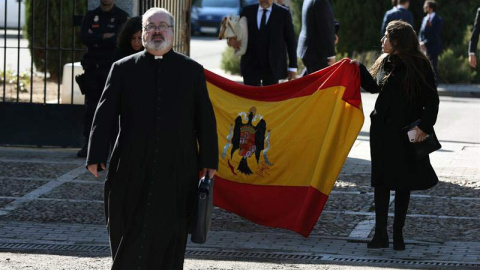 El sacerdote Ramón Tejero, que ha oficiado la misa de la reinhumación de Francisco Franco, a su salida del cementerio de Mingorrubio junto a dos familiares del dictador que portan una bandera española con el escudo franquista. (JAVIER LIZÓN | EFE)