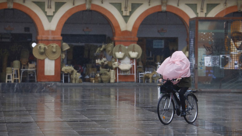 Una mujer en bicicleta a la que el viento le ha tapado la cara totalmente con el chubasquero mientras pasa por la Plaza de la Corredera de Córdoba Una mujer en bicicleta a la que el viento le ha tapado la cara totalmente con el chubasquero mientras pasa por la Plaza de la Corredera de Córdoba