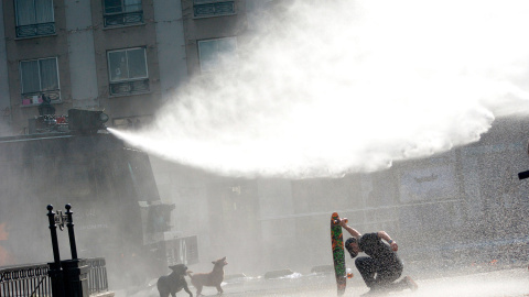 Un manifestante se protege con su monopatín de un chorro de agua a presión lanzado por los antidisturbios durante una manifestación para exigir cambios en el sistema educativo de Santiago (Chile).- CATALINA ALLEN (REUTERS)