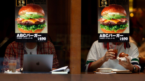 Dos clientes dentro de una tienda de comida rápida, tras sendos carteles de hamburguesas, en un restaurante de comida rápida en un distrito comercial de Tokio (Japón).- YUYA SHINO (REUTERS)