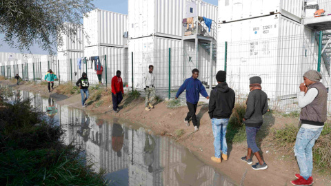 Varios inmigrantes se reflejan en el agua fangosa al pasar junto a los contenedores utilizados como vivienda improvisada, en la víspera del   desmantelamiento de La Jungla de Calais.- PHILIPPE WOJAZER (REUTERS)