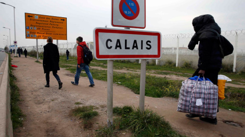 Un inmigrante traslada bosas con sus pertenencias junto a la señal que indica el límite urbano de la ciudad francesa de Calais.- PASCAL   ROSSIGNOL (REUTERS)