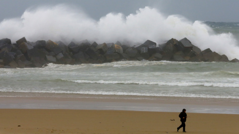 Un hombre se protege contra el viento y la lluvia en la playa de la Zurriola de San Sebastián, donde los cielos permanecen cubiertos con lluvias y chubascos generalizados. EFE/Juan Herrero.