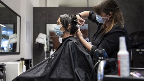 27/04/2020.- Una peluquera corta el pelo a una mujer con una máscara facial protectora contra el coronavirus en una peluquería de Berna, Suiza este lunes. Servicios como la peluquería, los masajes, los tatuajes o los salones de belleza está