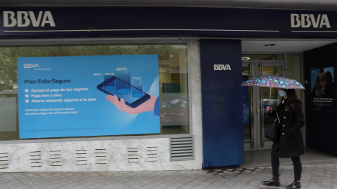 Una mujer con paraguas pasa por delante de la puerta de una sucursal de BBVA en Madrid. E.P./Isabel Infantes