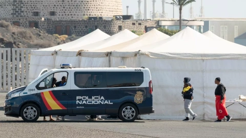 Tres mujeres pasan ante un coche de policía y las carpas del campamento provisional en Arrecife.