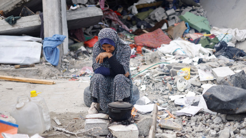 Una mujer palestina está sentada junto a un edificio mientras espera a que se caliente una cacerola.