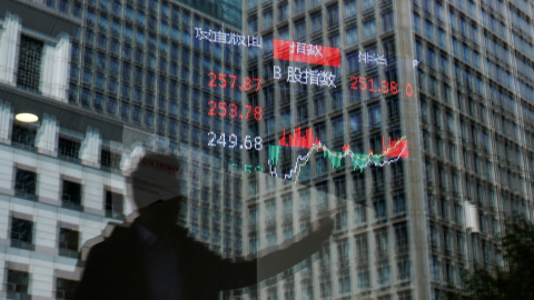 Una persona y varios edificios se reflejan en el cristal de una casa de bolsa en cuyo interior hay colgado un tablón de anuncios con información sobre el índice bursátil, en Pekín, China, el 9 de abril de 2025.