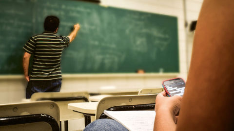 Una alumno, en clase con el móvil mientras su profesor imparte la lección.